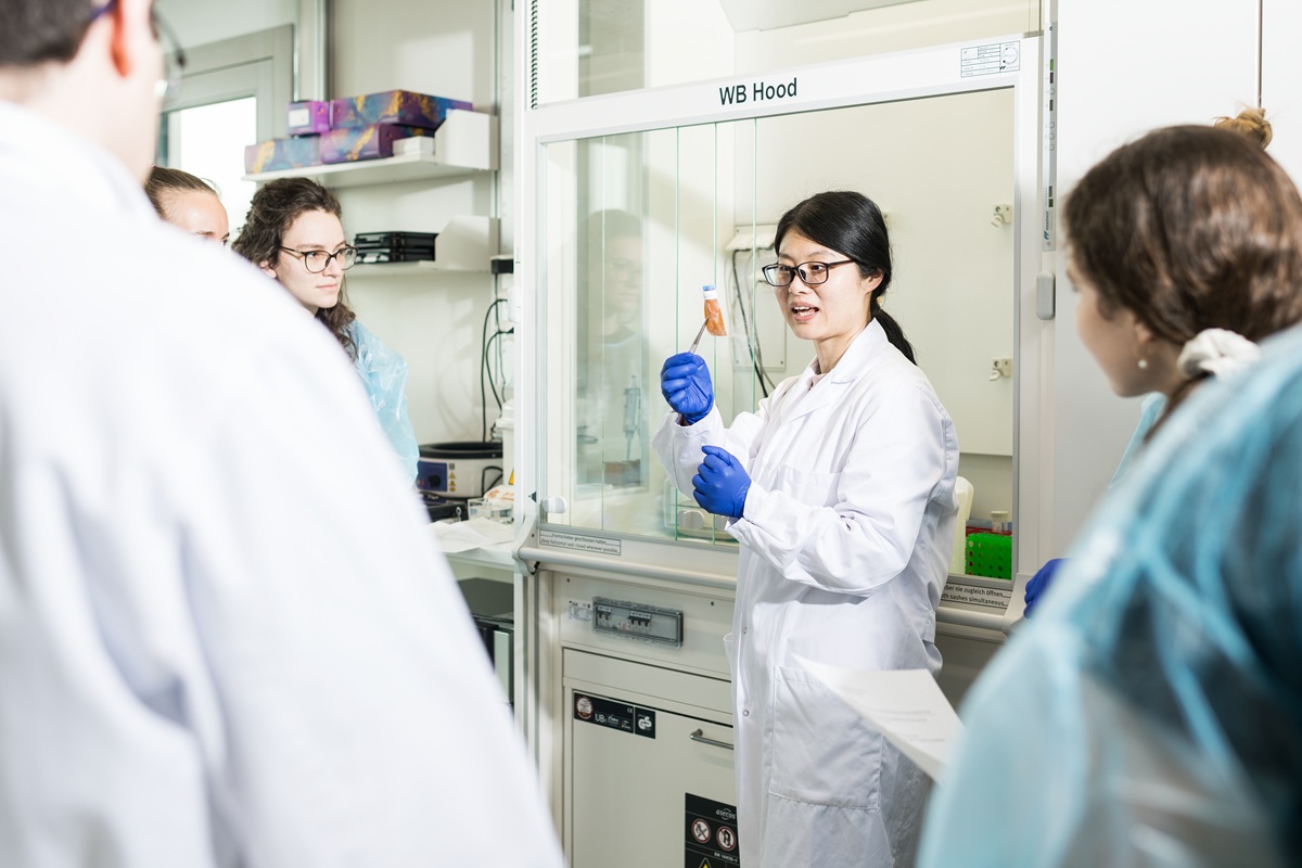 Researcher in a laboratory wearing a white lab coat and blue gloves holding a sample tube while explaining it to colleagues near a laboratory hood.