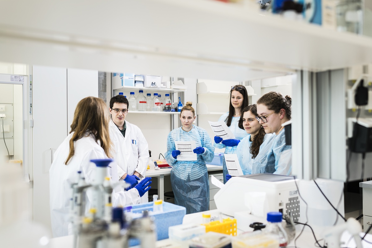 Scientist in a lab coat explaining something to colleagues beside laboratory equipment.